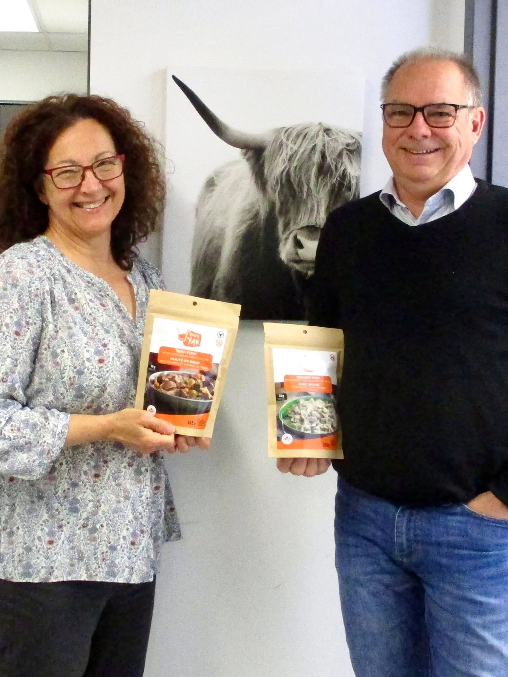 Christine Chénard and Guy DuBuc, co-owners of Happy Yak, pose inside in front of a wall, each holding a packet of freeze-dried food. A photo of a yak hangs on the wall in the background.